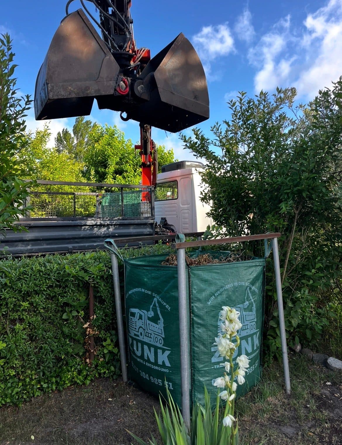 Greifer-Entsorgung eines Big Bags Ladekran-Greifer hebt einen grünen, mit Grünschnitt gefüllten Big Bag von Zunk Transporte und Entsorgung über einen Gartenzaun