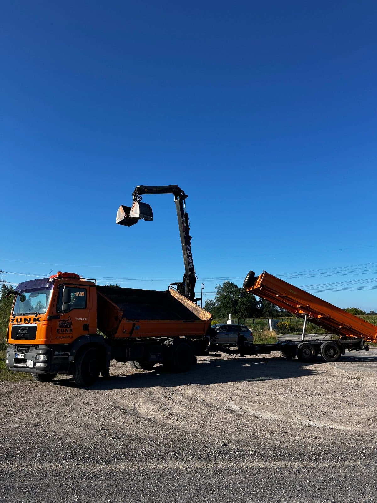 Zunk-Fahrzeug mit Kipper-Anhänger Oranger Zunk-LKW mit ausgefahrenem Ladekran und angehobenem Kipper-Anhänger vor blauem Himmel